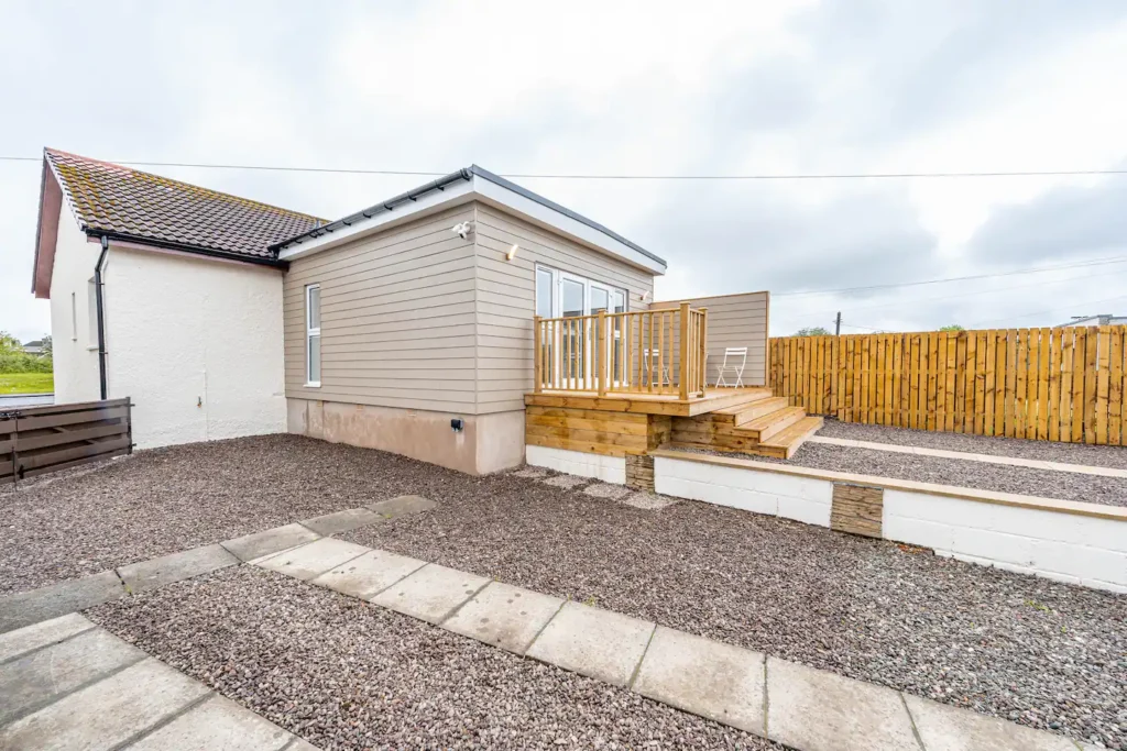 Modern house with wooden deck and gravel yard, fenced by timber panels under a cloudy sky.