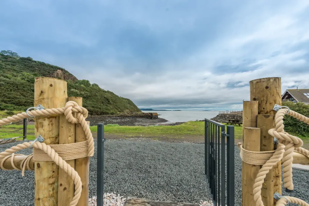 Coastal view with wooden posts and ropes, overlooking lush greenery and a serene ocean under a cloudy sky.