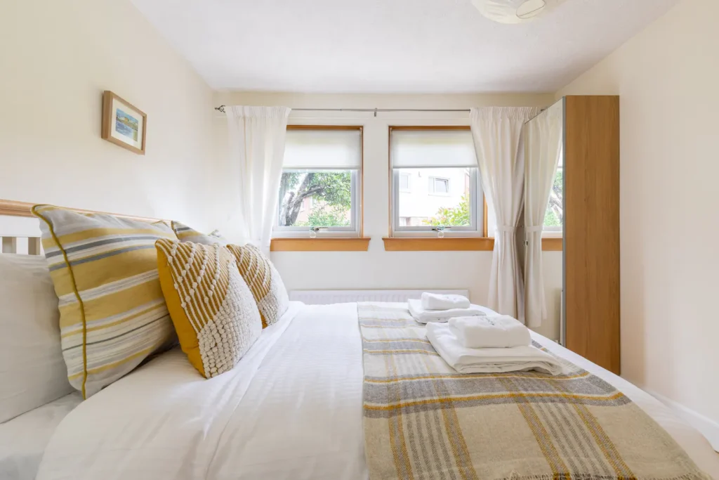 Cozy bedroom with striped pillows, fresh linens, and natural light from two windows with white curtains.