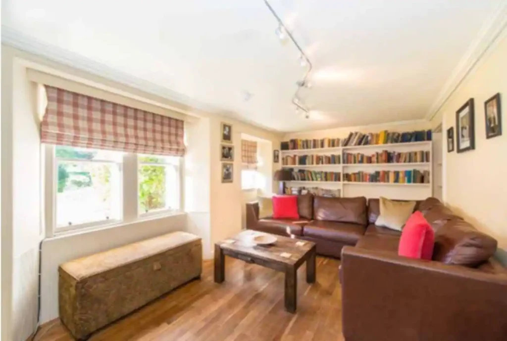 Cozy living room with brown leather sofa, wooden coffee table, bookshelves, and plaid window blinds.