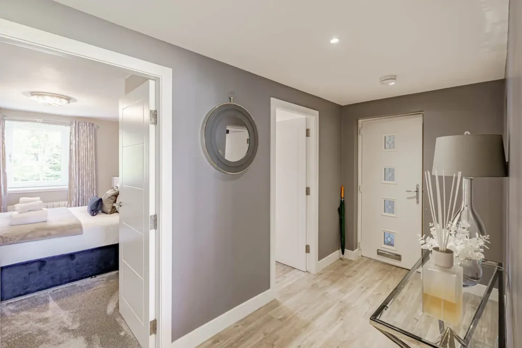 Modern hallway with grey walls, decorative mirror, and vase on table; open door to a cozy bedroom with natural light.