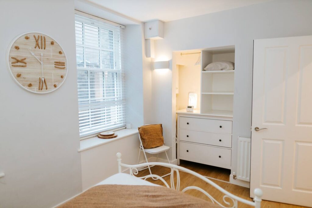 Cozy bedroom with wooden clock, white dresser, and window with blinds, offering a bright and minimalist design.