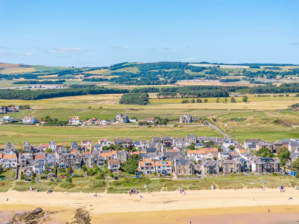 Aerial view of coastal town with sandy beach and green countryside under a clear blue sky.