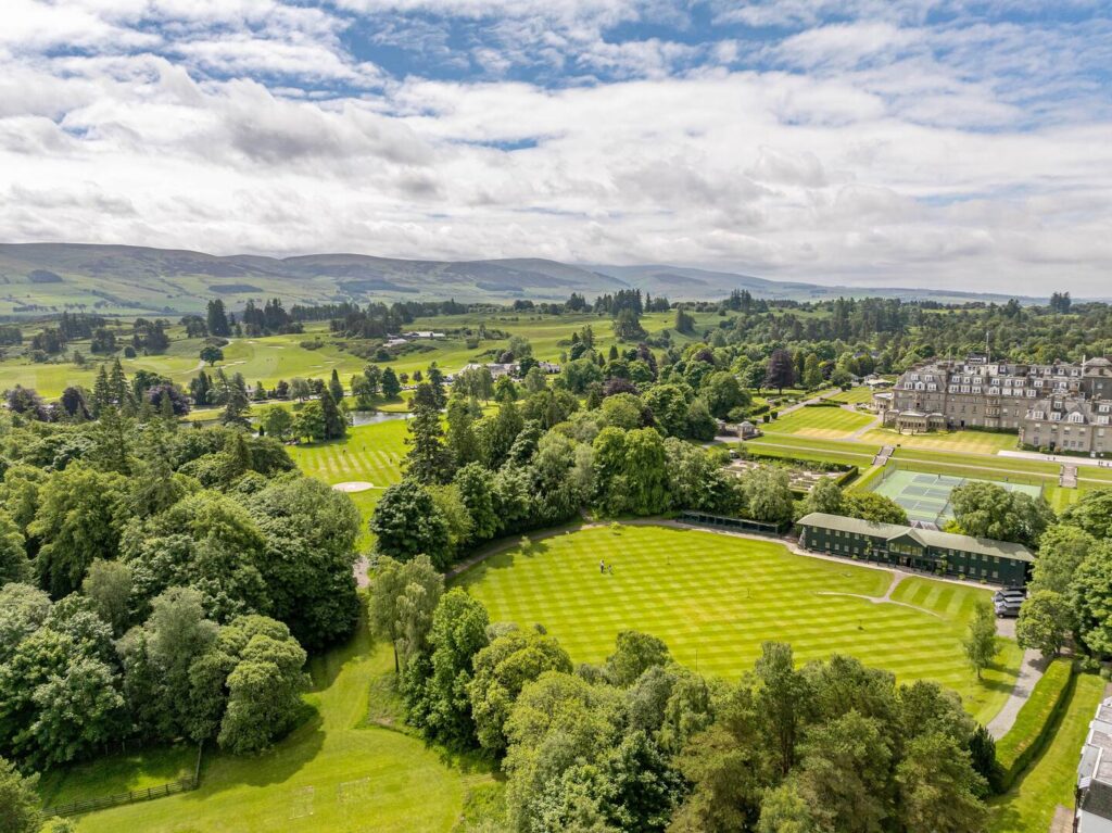 Aerial view of lush green parkland, tennis courts, and historic buildings under a cloudy sky.