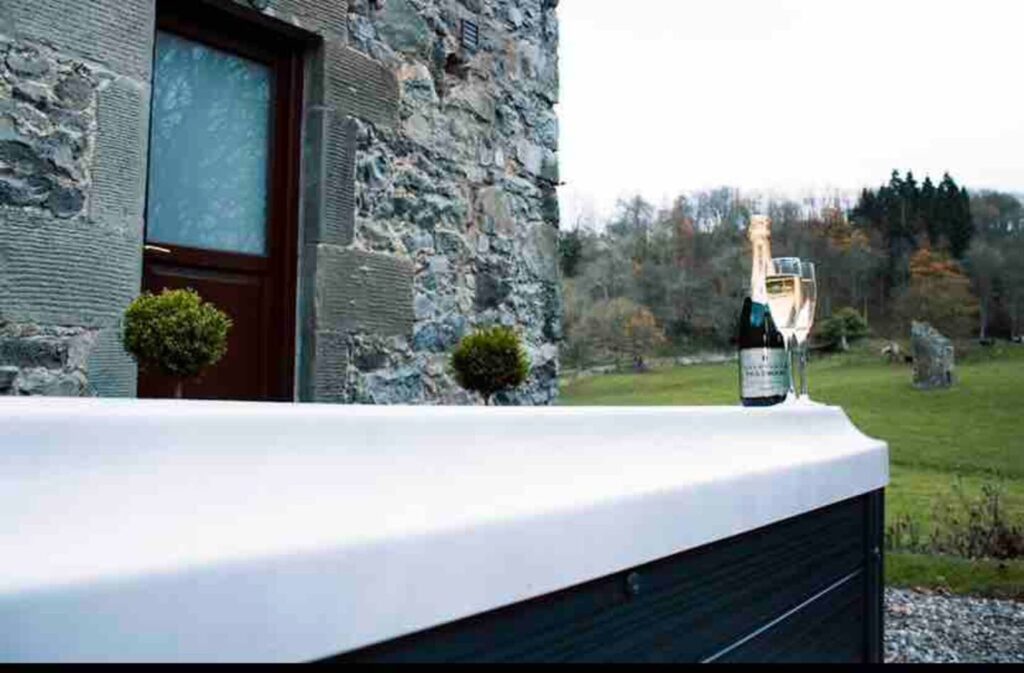 Bottle and glass of champagne on a hot tub outside a rustic stone building with countryside views.