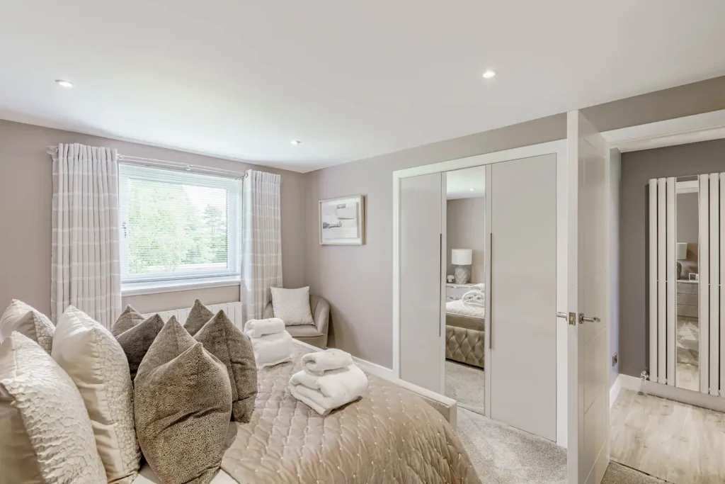 Modern bedroom with plush pillows, neatly folded towels, large windows, and mirrored wardrobe reflecting cozy decor.