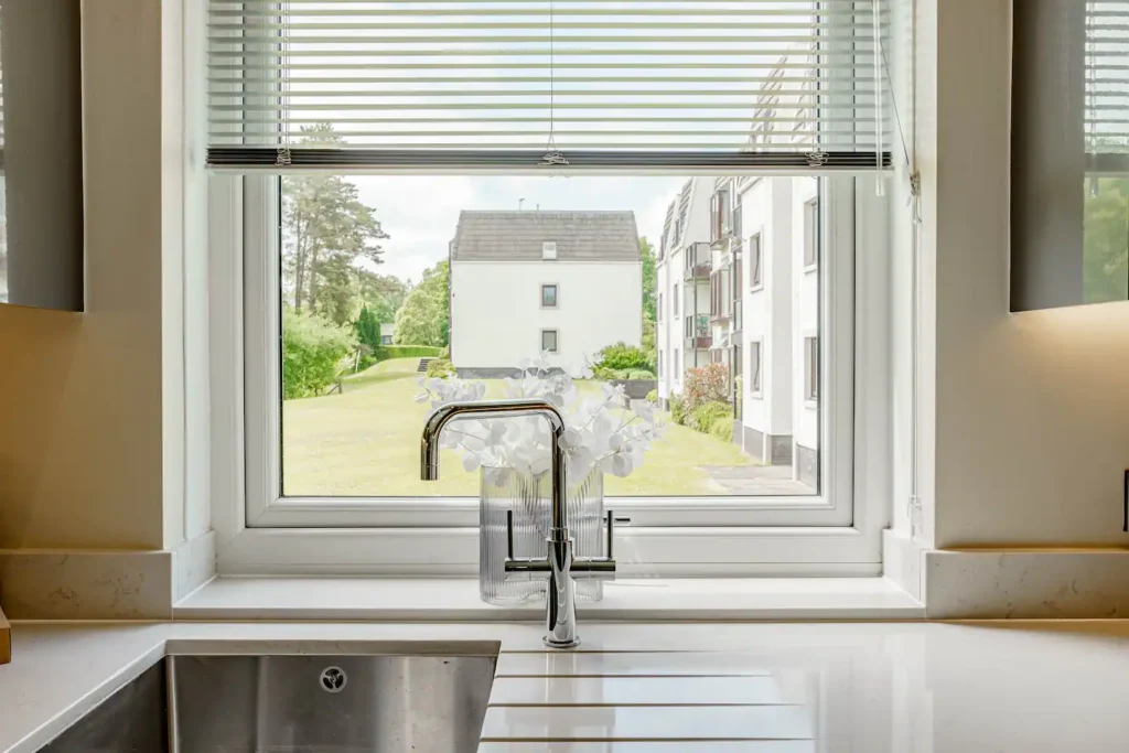 Modern kitchen sink with a vase of flowers by a window overlooking a green lawn and buildings.