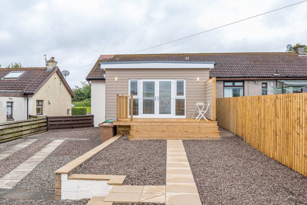 Modern bungalow with wooden deck and gravel pathway, featuring a small outdoor table and chair set.