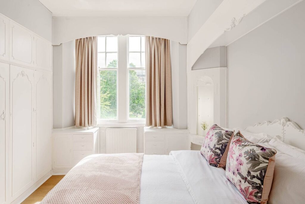 Bright, cozy bedroom with floral pillows, soft beige curtains, and white furniture by large windows.