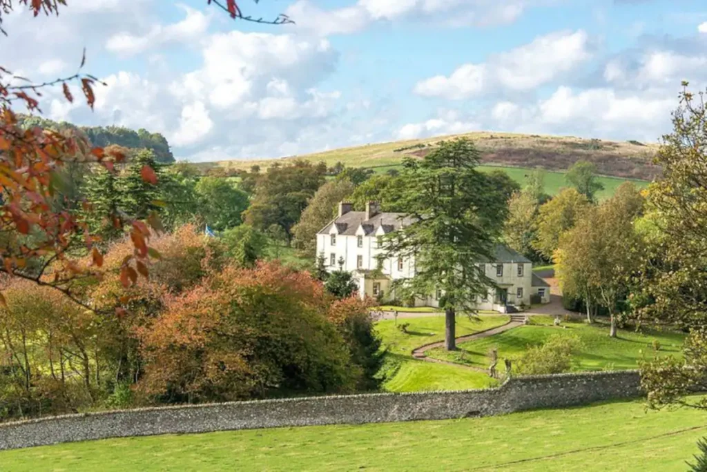 Charming countryside mansion surrounded by lush greenery and scenic hills under a bright blue sky.