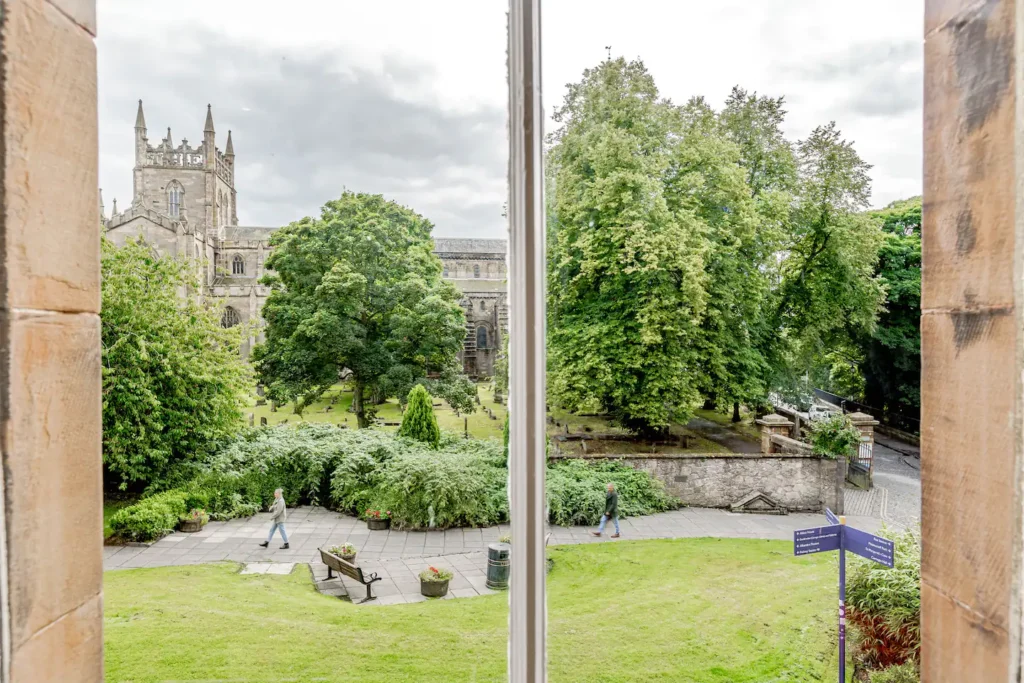 View of lush green garden and historic stone building from a window, with two people walking on a path.