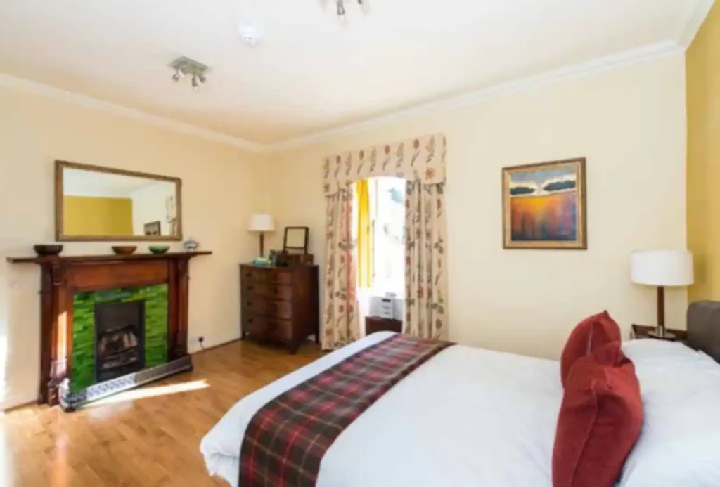 Cozy bedroom with wooden fireplace, plaid bedspread, and framed art, filled with natural light.