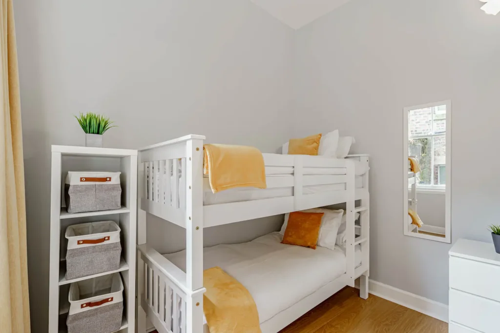 Modern bedroom with white bunk beds, orange accents, storage baskets, and a mirror reflecting natural light.