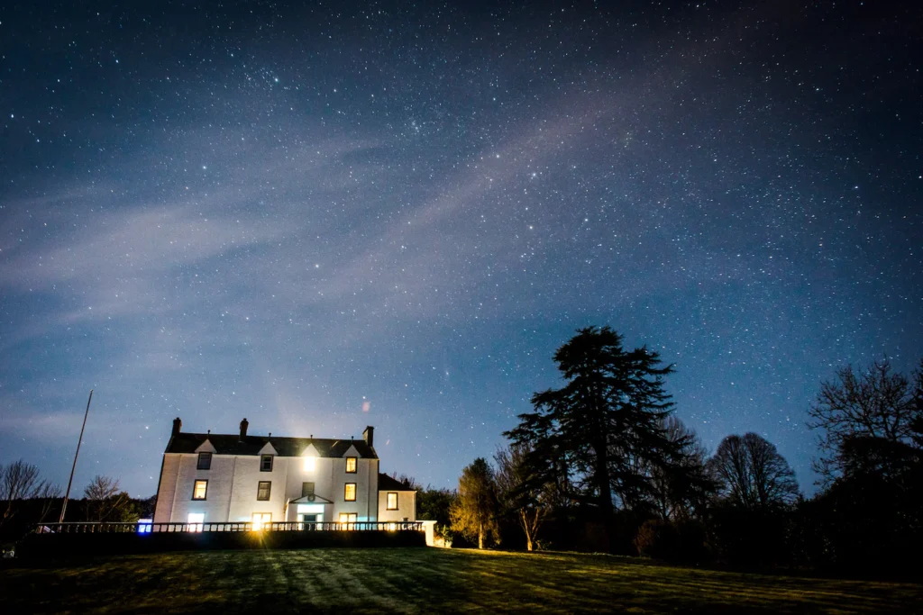 Starry night sky over illuminated house and trees, showcasing serene countryside landscape.