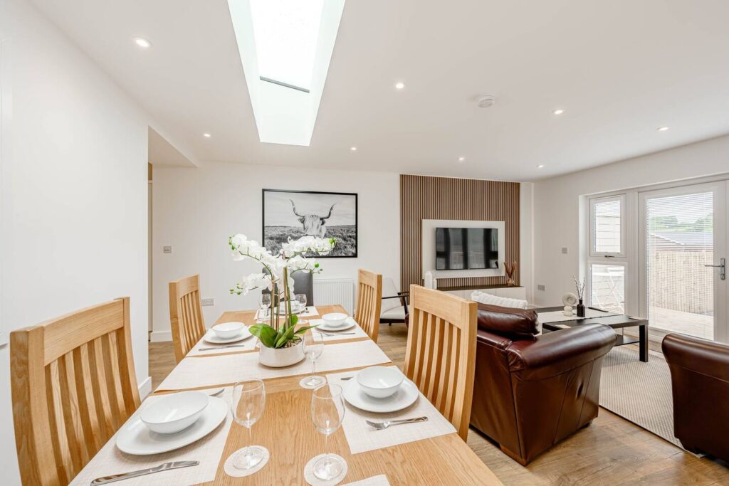 Modern dining and living room with wooden table, leather sofas, and large window, featuring a skylight and plant decor.