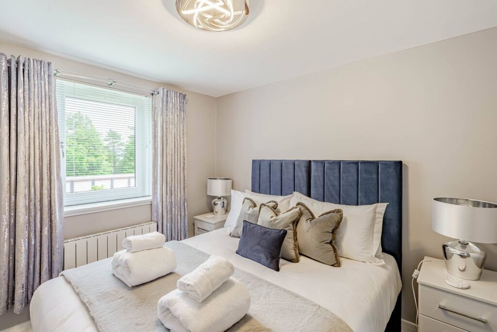 Modern bedroom with a plush bed, decorative pillows, fresh towels, and natural light through large window.