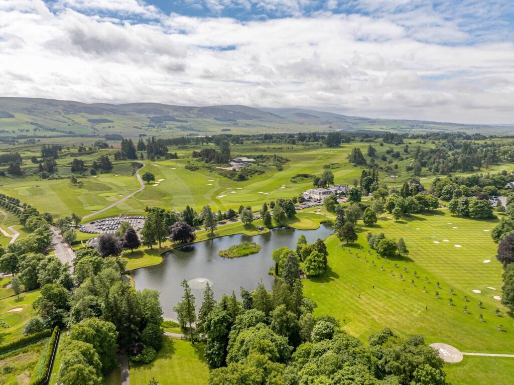 Aerial view of a lush golf course with rolling hills, trees, and a serene pond on a sunny day.