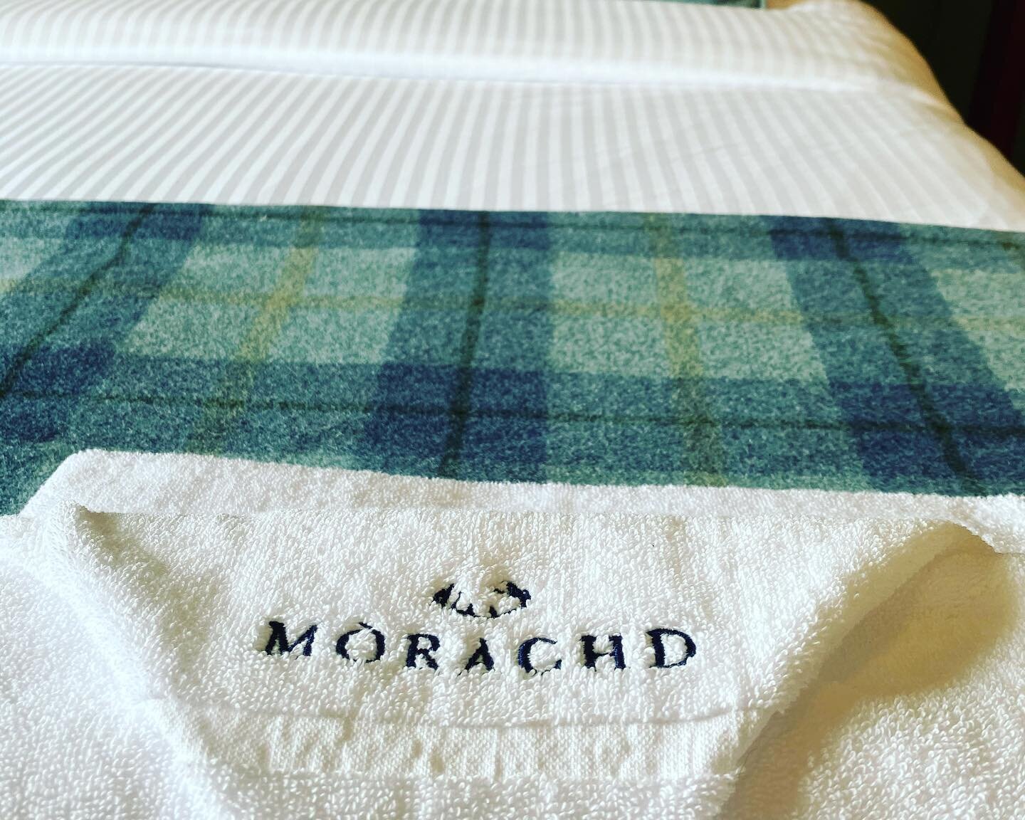 Close-up of a hotel bed with striped sheets and a plaid blanket, featuring a towel embroidered with Mòrachd text.