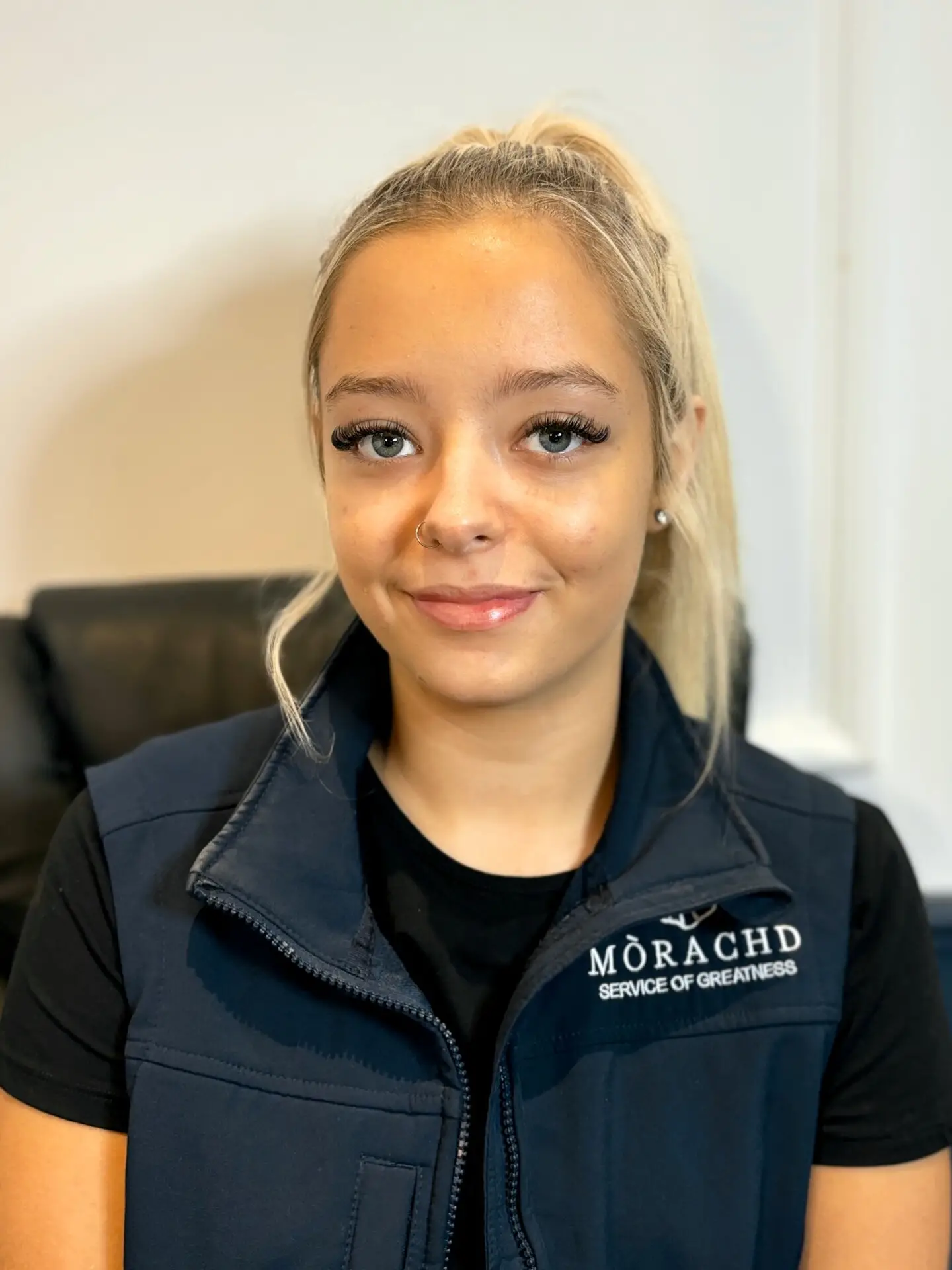 Young woman in a navy vest with Mòrachd Service of Greatness logo, smiling confidently indoors.