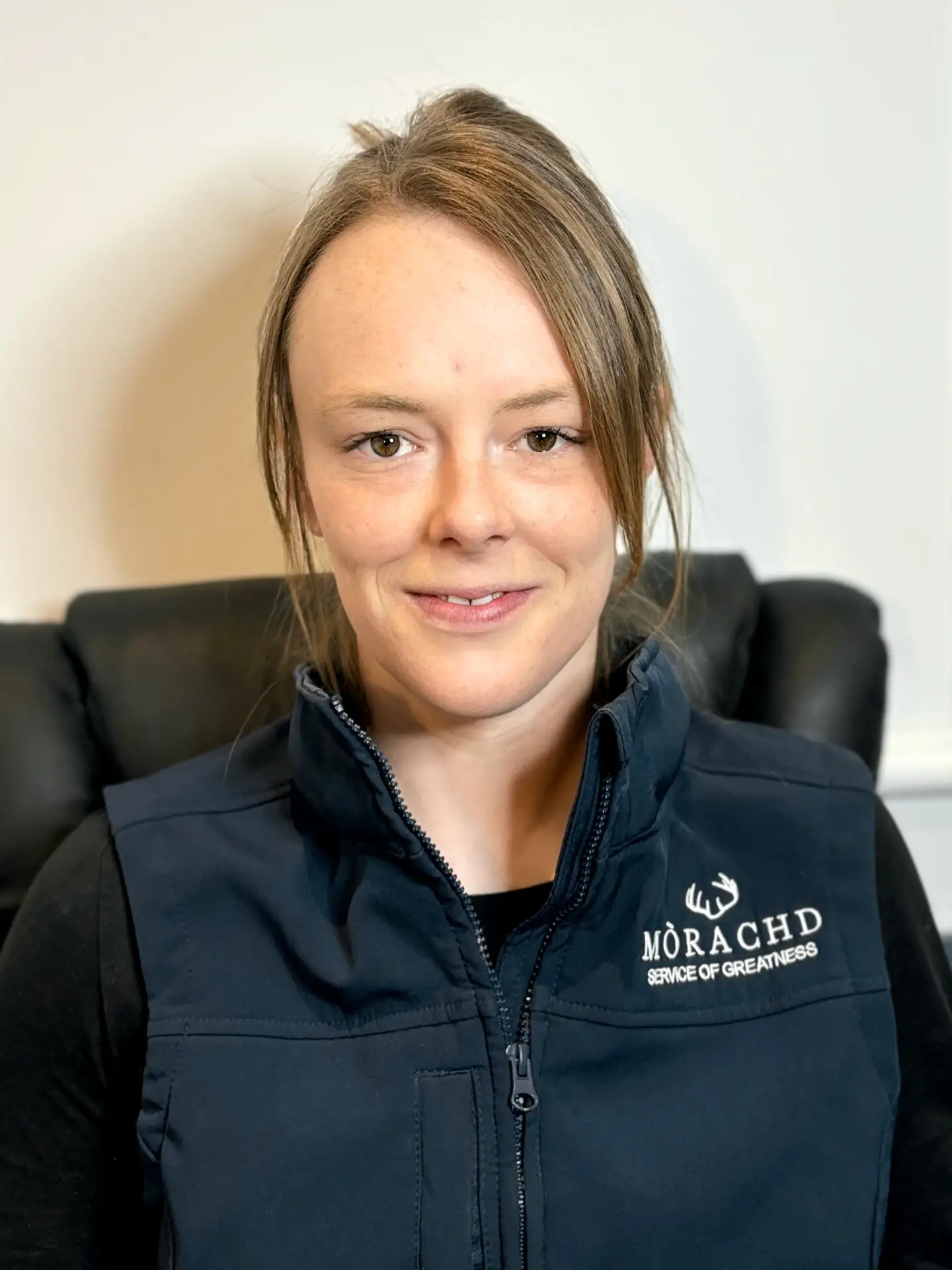 Smiling person in a navy vest with Mòrachd Service of Greatness logo, seated in an office chair.