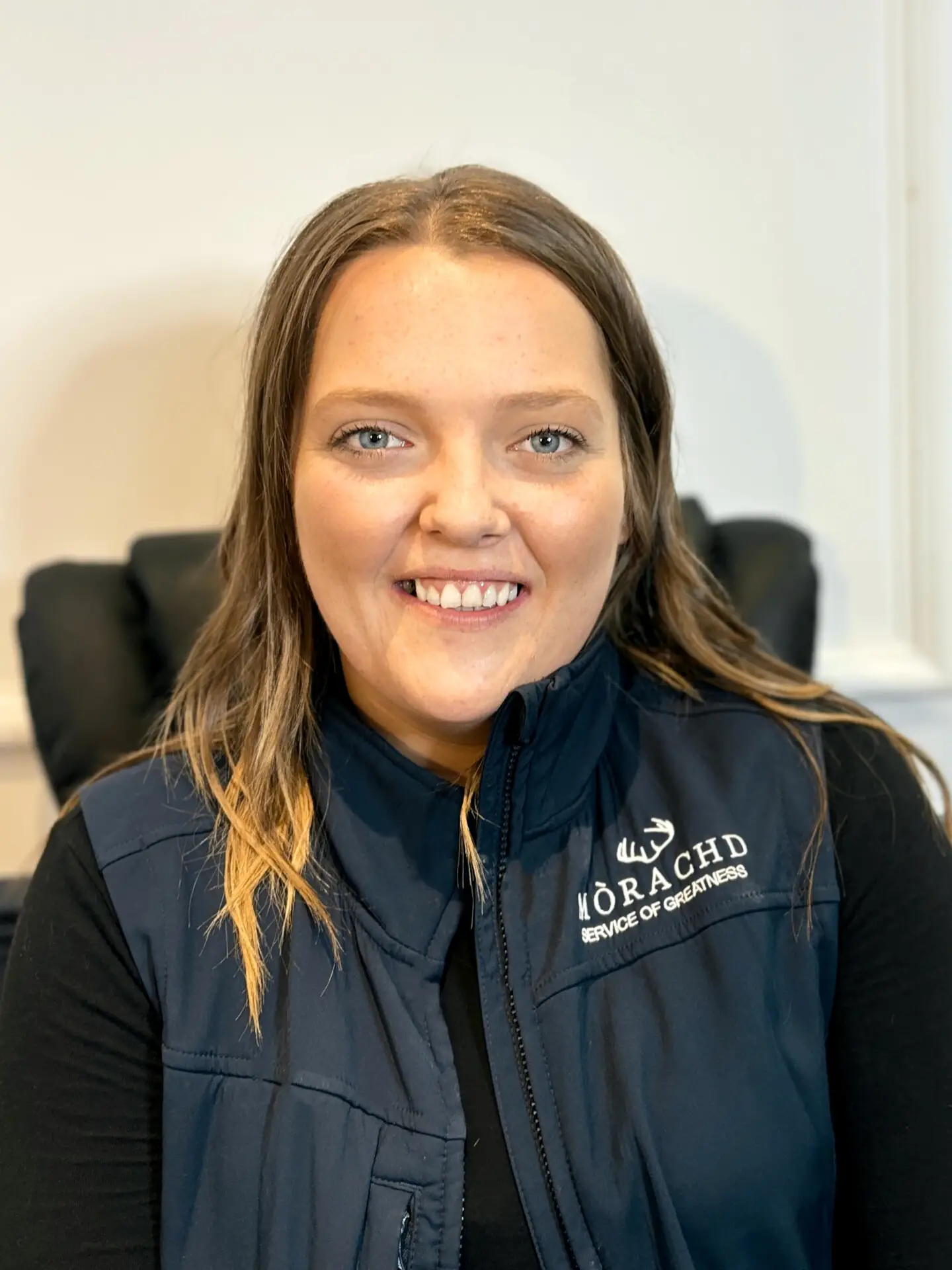 Smiling woman wearing a Mòrachd Service of Greatness jacket, sitting in an office setting.