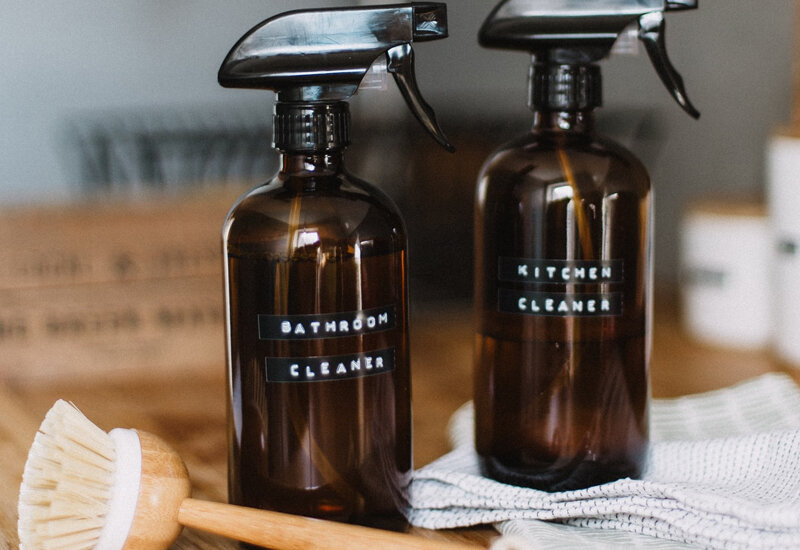 Amber glass spray bottles labeled bathroom and kitchen cleaner on a wooden surface with a scrub brush.