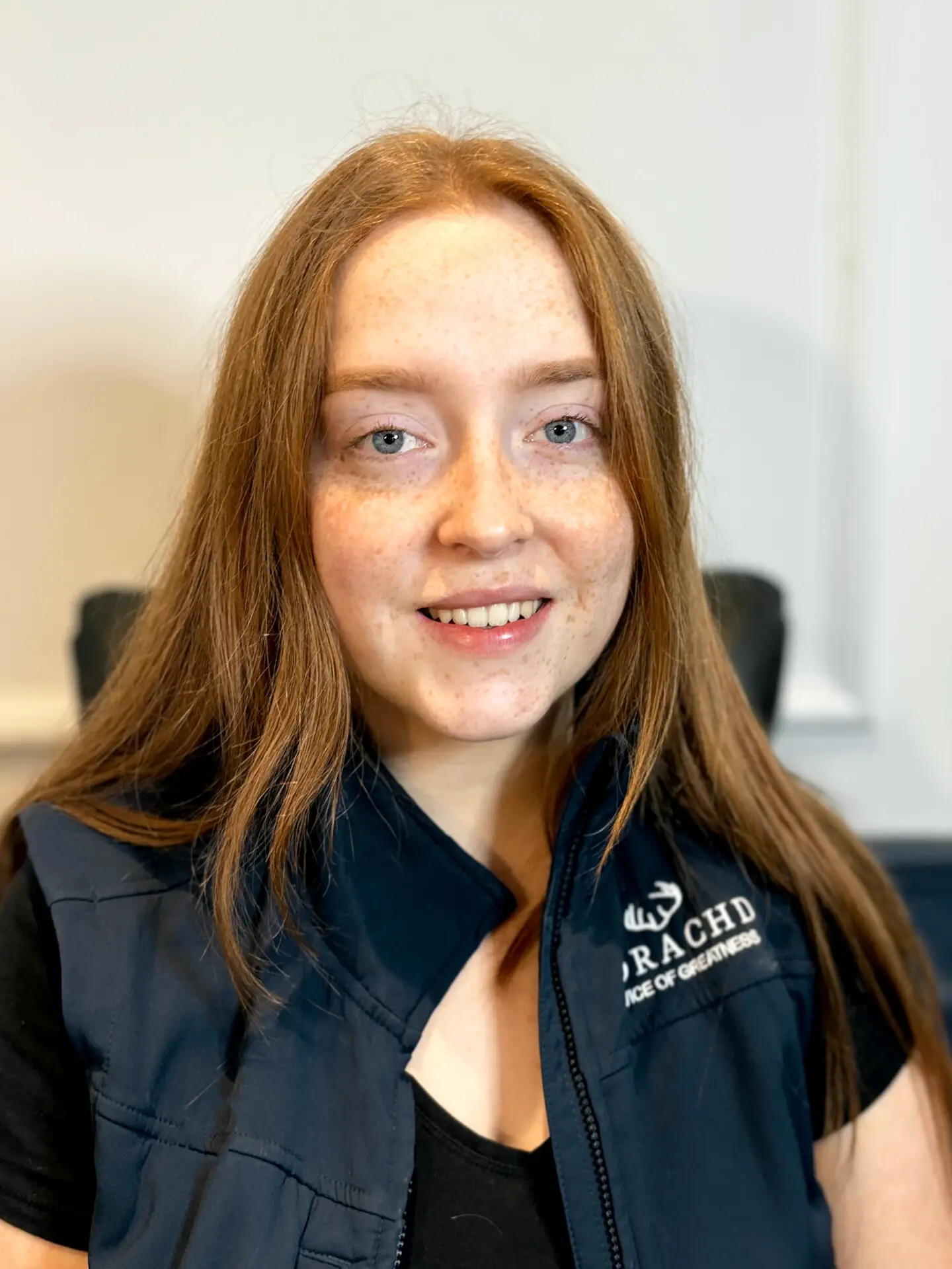 Smiling person in a branded vest, seated indoors with natural lighting.