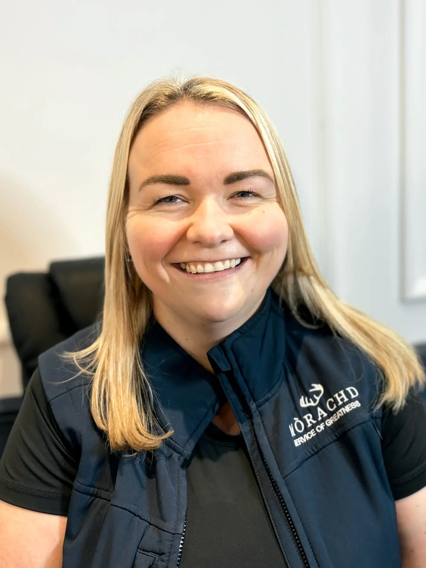 Smiling woman in a black branded vest seated indoors.