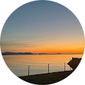 Sunset view over calm waters with a silhouette of a fence in the foreground and vibrant orange and blue sky.