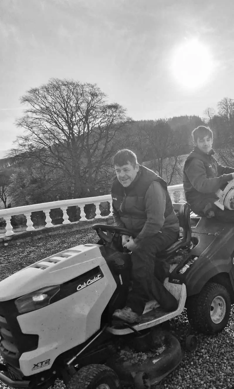 Two men on a riding lawnmower with a scenic background of trees and the sun.