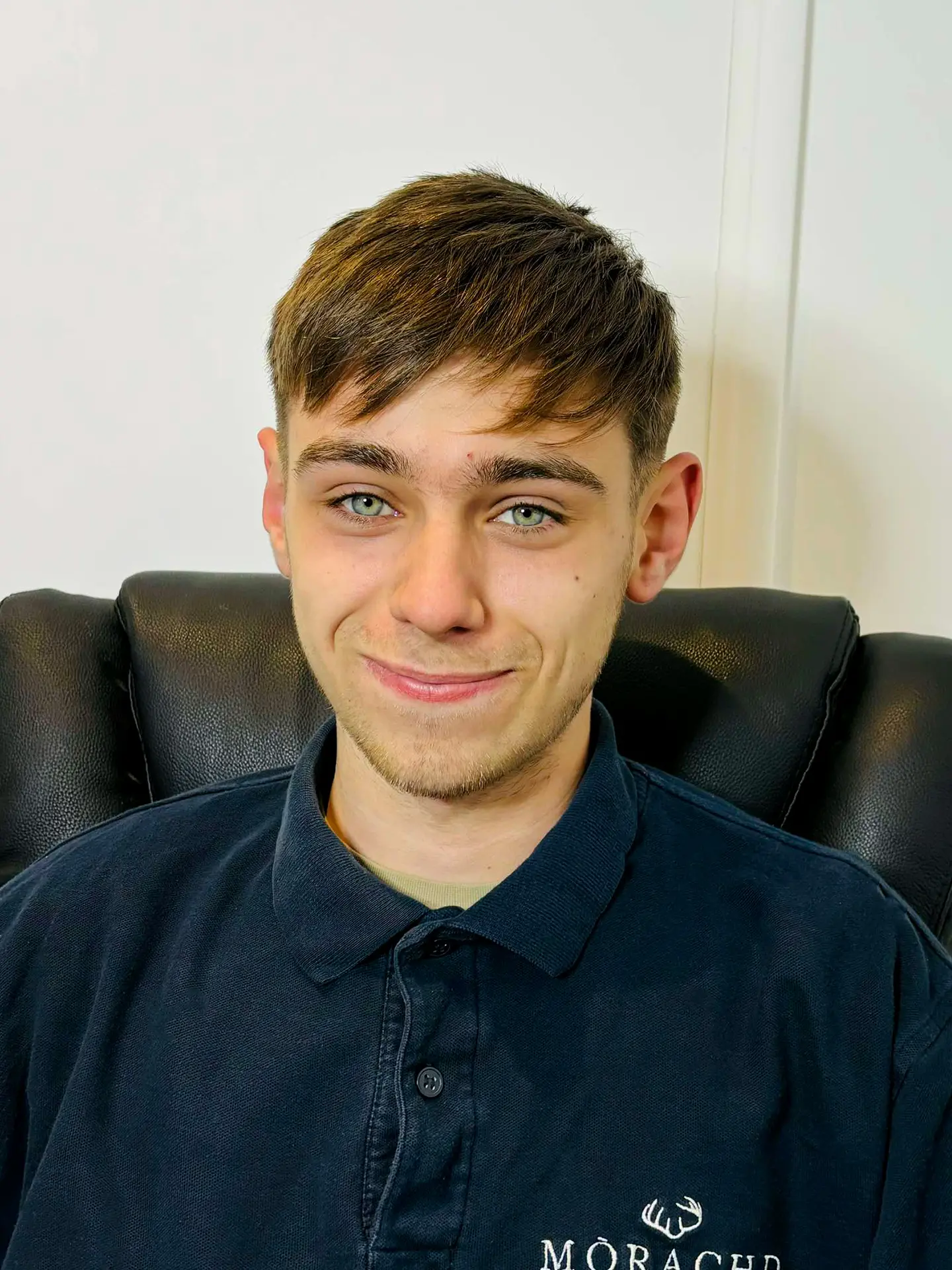 Smiling young man in a black polo shirt seated on a leather chair.