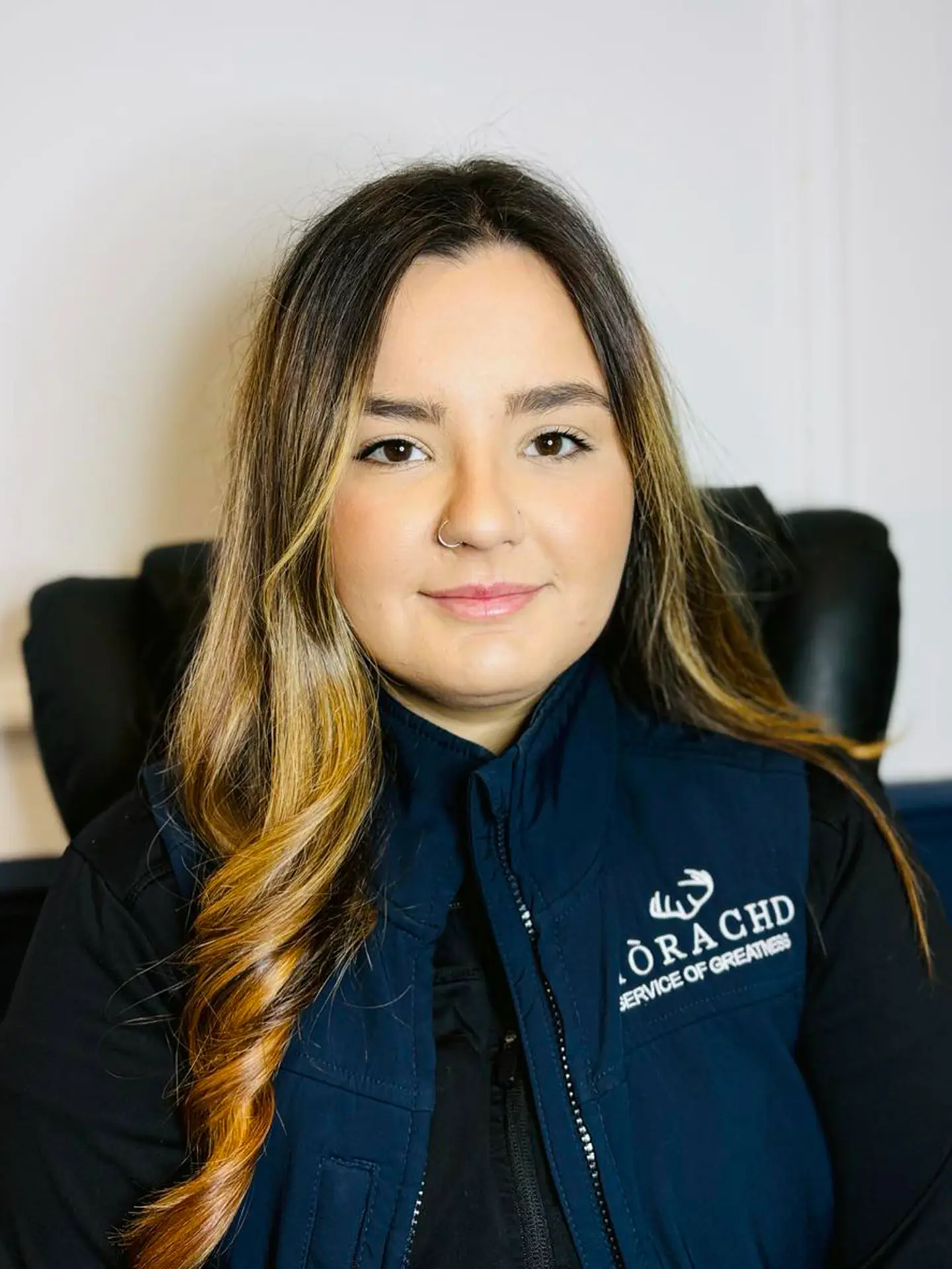 Portrait of a woman with long hair, wearing a blue vest, seated in a black chair.